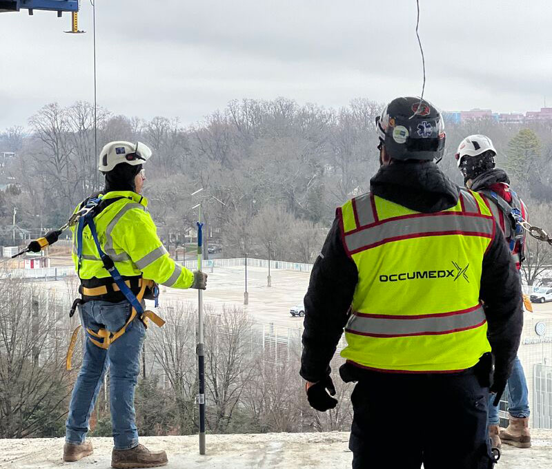 OccuMedX medic in a safety vest observing a worker in a fall protection harness on a construction site